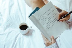 A woman sitting on a bed, focused on writing in a notebook with a pen in her hand.