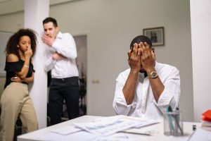 A man standing in front of a desk, hiding his face with his hands in frustration or distress.