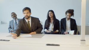 A diverse group of people sitting at a table, engaged in a discussion, with a well-dressed man in a suit leading the conversation.