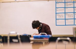 A student writing on a whiteboard in a classroom, surrounded by colorful educational posters.