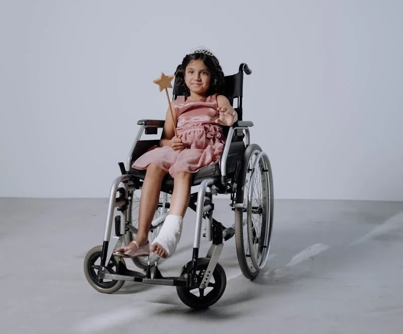 a little girl in a pink dress seated on a wheelchair with an injured foot